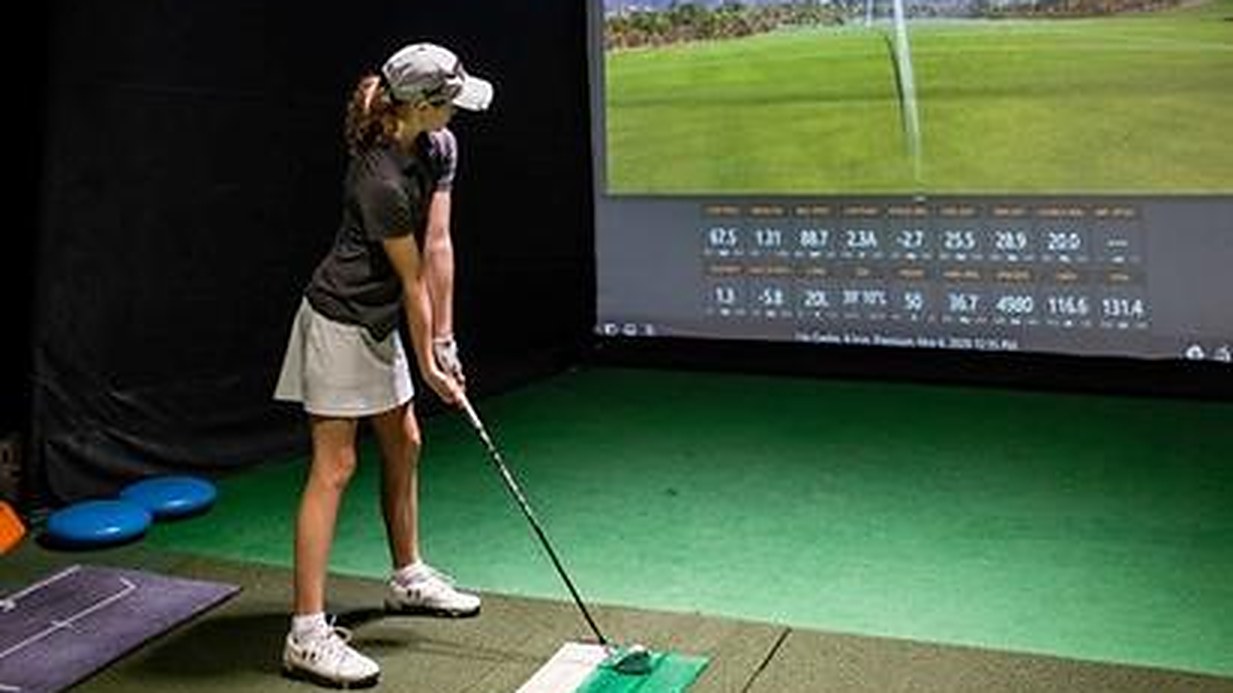 A golfer mid-swing in front of a TrackMan simulator screen at Oakmont Golf Center